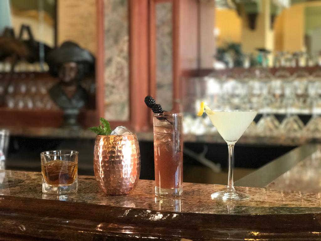 Three cocktails sit on a polished bar top: a whiskey glass, a copper mug filled with a fruity drink, and a sleek martini with a lemon twist, showcasing the bar's offerings.